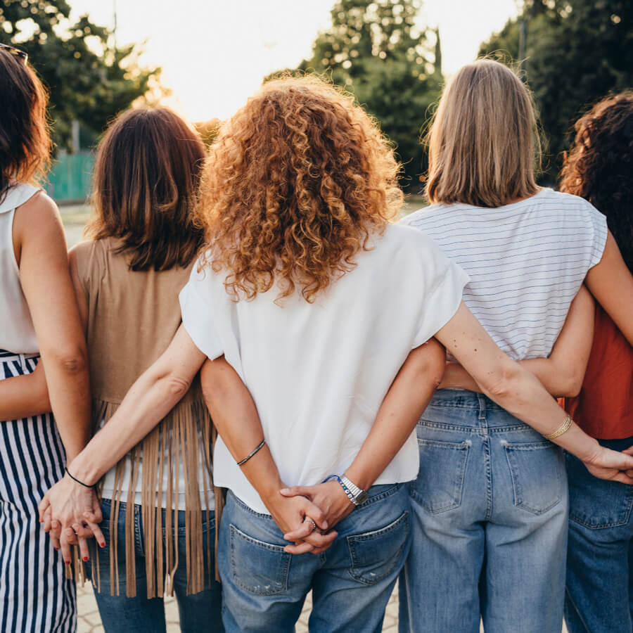 Five women standing arm in arm outdoors, photographed from behind